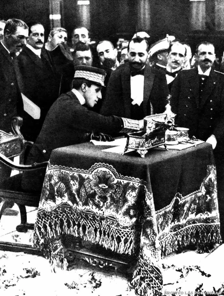King Alfonso XIII signing at a cloth-covered table, surrounded by dignitaries in frock coats and hats
