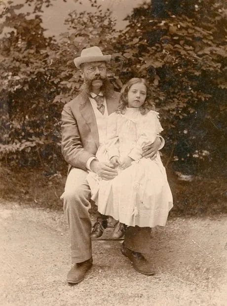 Fotografía sepia de un caballero con sombrero sentado en un jardín con una niña en el regazo