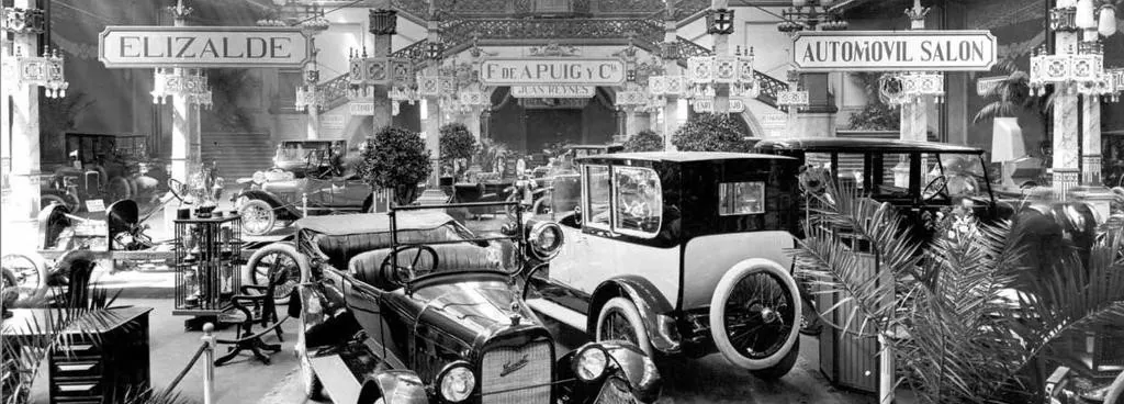 Salón de exposición de automóviles de principios del siglo XX con muchos coches antiguos y rótulos de marcas